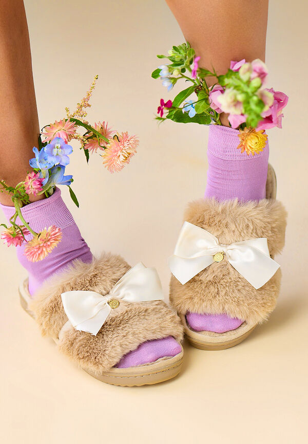 Person wearing tan faux fur slip-on slippers with purple socks, flowers stuffed into the socks.