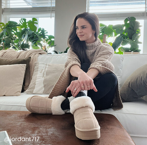 A woman sits on a white sofa with her legs crossed, wearing a beige sweater, black pants, and light tan slippers, with green plants and pillows in the background.