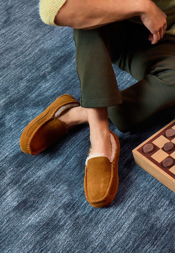 A man wearing green pants and brown moccasin slippers sits on a carpeted floor near a checkers board.