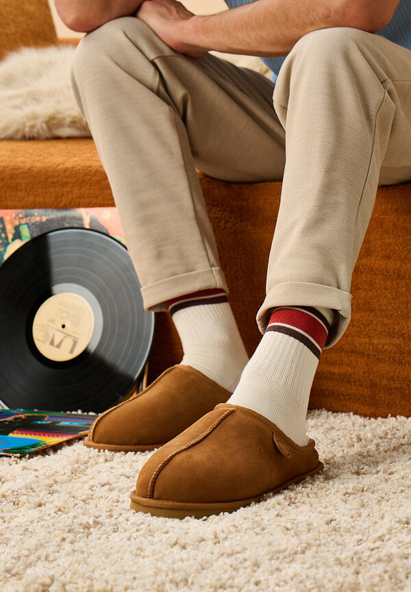 A man wearing beige pants, white socks with stripes, and brown slippers sits on a carpeted floor near a vinyl record and a brown couch.