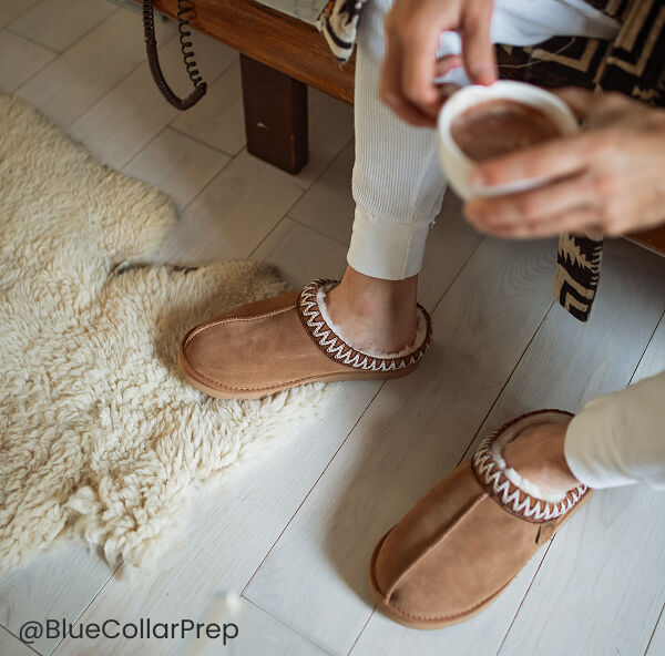 Man wearing tan men's shearling clogs and white pants sits on a light wood floor next to a cream rug, holding a cup.