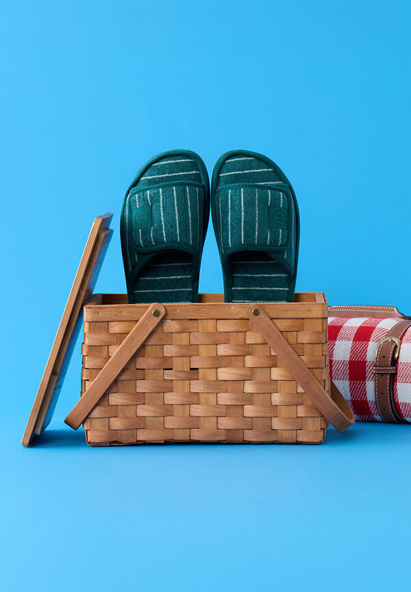 Dark teal striped slide slippers displayed inside an open wicker picnic basket against a bright blue background.