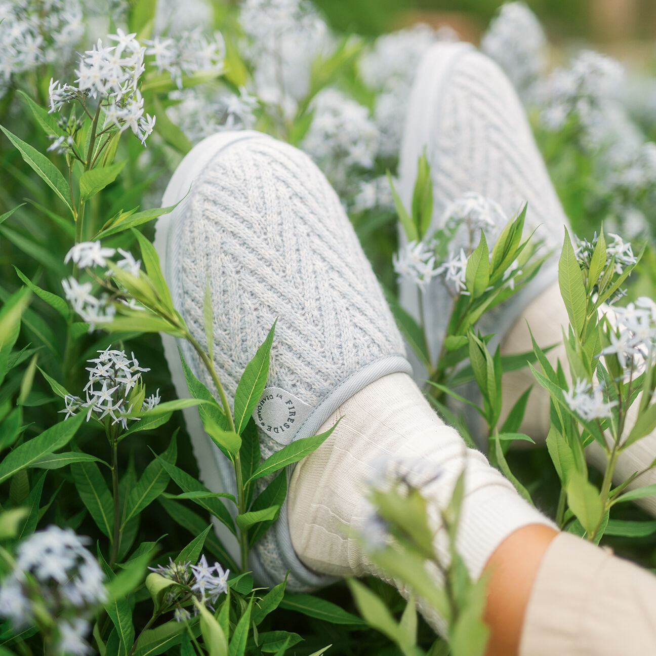 Blue knit house slippers worn with cream socks, surrounded by green leaves and small white flowers outdoors.