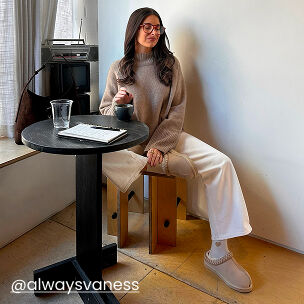 Woman seated at a caf&eacute; table wearing beige slippers with a neutral sweater and wide-leg pants.
