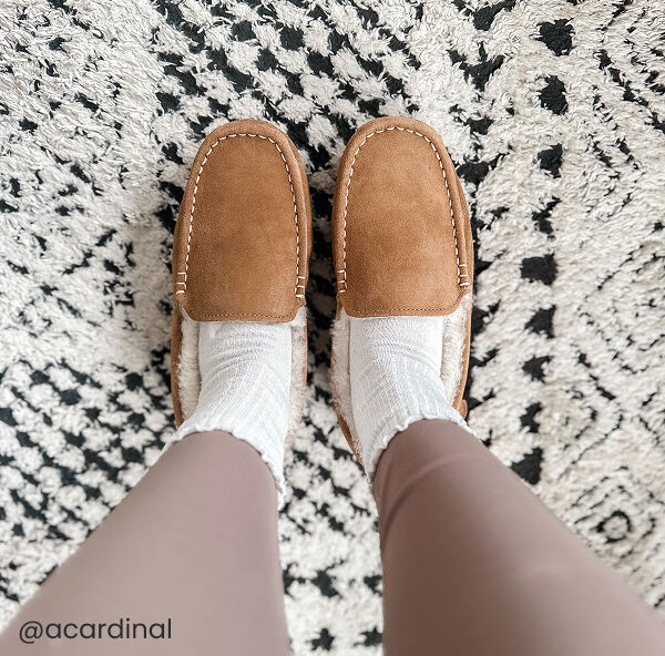 A woman wearing white socks and brown suede slippers stands on a black and white patterned rug.