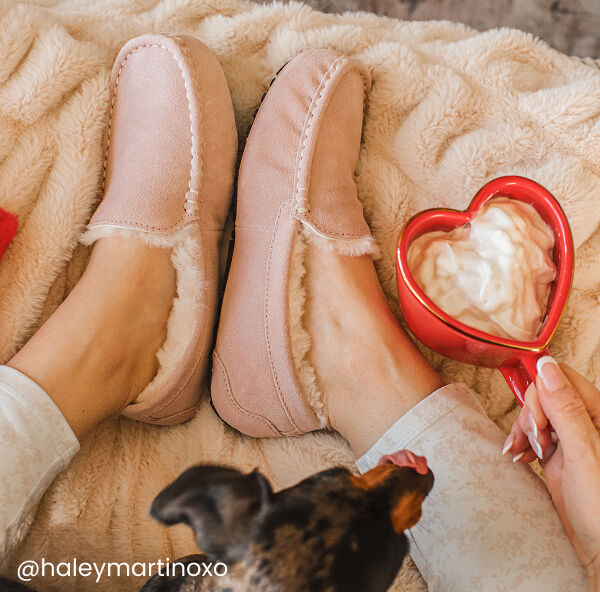 Woman wearing pink slippers sits on a fluffy blanket, holding a red heart-shaped mug with a creamy drink; a small dog is visible in the lower part of the image.