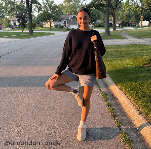 A woman stands on a suburban street at sunset, balancing on one leg with the other foot against her knee, wearing a black sweater and, gray shorts, and clogs.