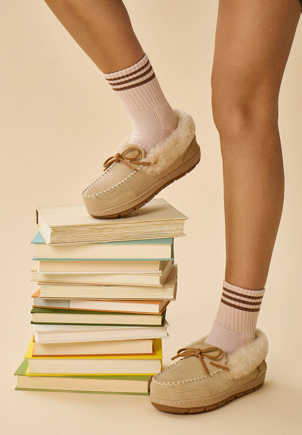 A woman wearing tan moccasin slippers and pink striped socks stands with one foot on a tall stack of books against a beige background.