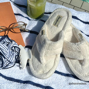 White plush terry flip flops resting on a striped towel beside sunglasses, a drink, and poolside accessories.