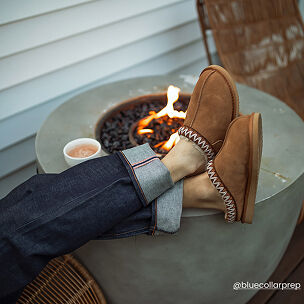 Person relaxing by an outdoor fire pit while wearing chestnut suede Fireside clogs with decorative whipstitch trim.