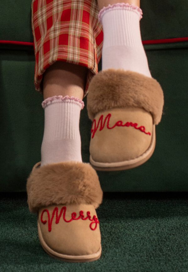 Tan faux-fur slippers embroidered &ldquo;Merry&rdquo; in red, worn with white ruffled socks.