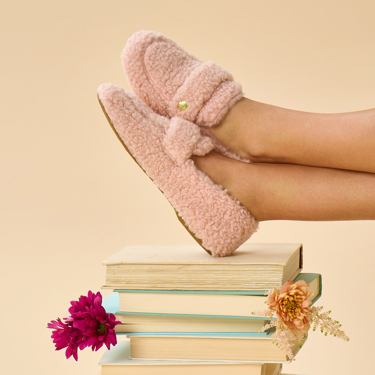 A pair of feet in pink fuzzy Dearfoam slippers rest on a stack of books, with pink and beige flowers tucked between the book pages.