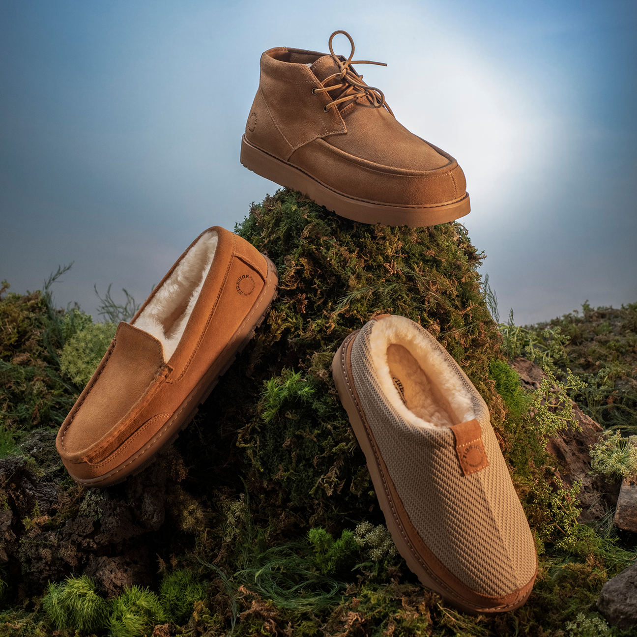 Three Dearfoams styles in warm brown tones, including a lace-up chukka, shearling-lined moccasin, and shearling-lined clog, displayed on a mossy outdoor landscape.