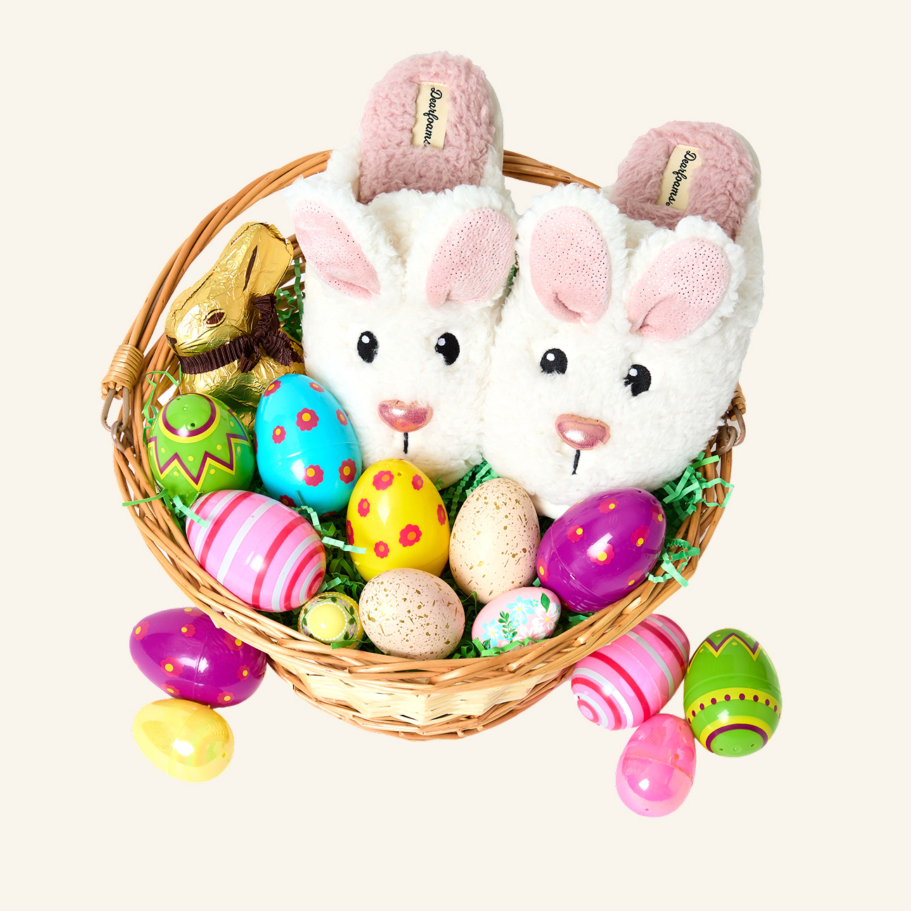 A wicker basket containing bunny slippers, a chocolate bunny, assorted decorated eggs, and colorful plastic eggs, sitting on a light background.