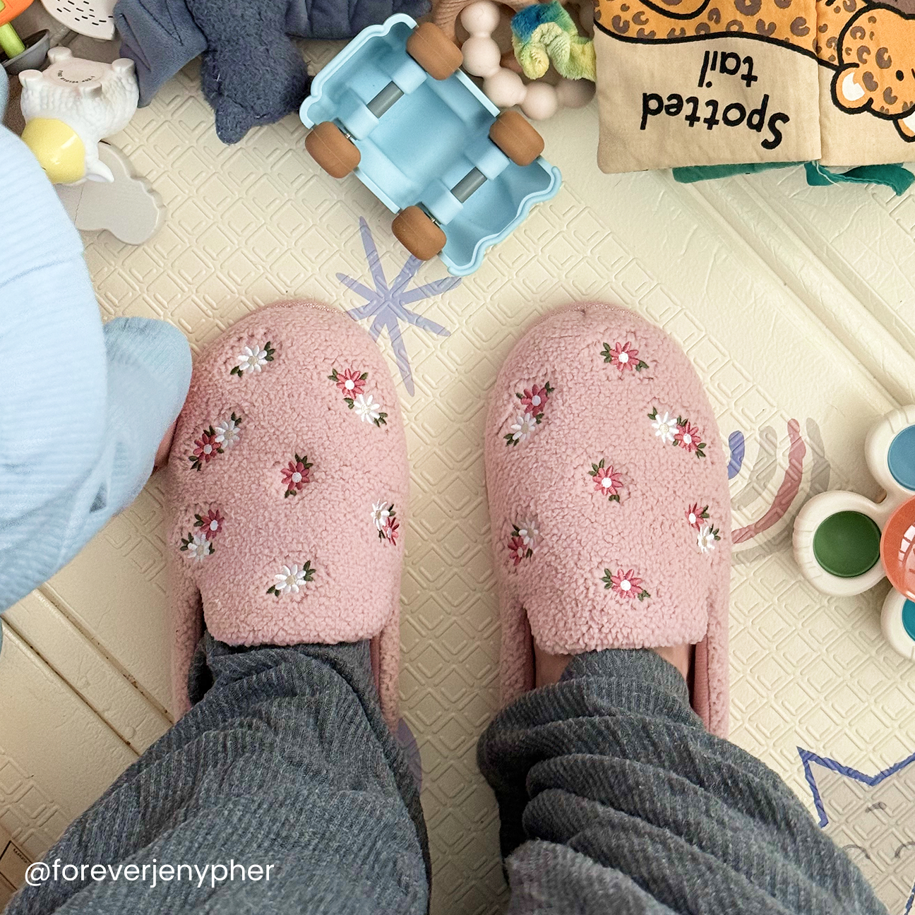 A mother wearing pink dearfoam slippers with floral designs stands on a patterned playmat surrounded by children's toys and a cloth book.