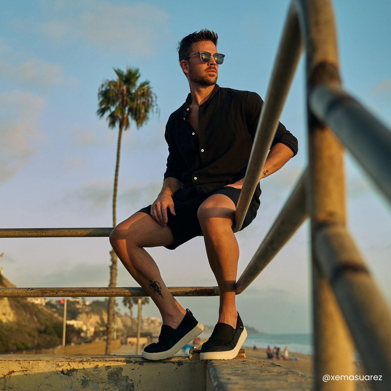 Man wearing black slip-on shoes with white soles while sitting on a railing near the beach.