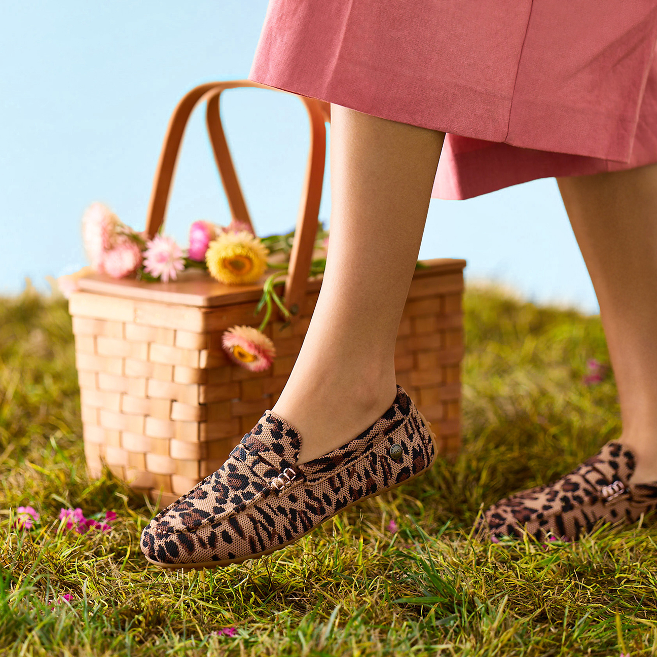 Woman wearing leopard print ballet flats outdoors beside a picnic basket in a grassy field.