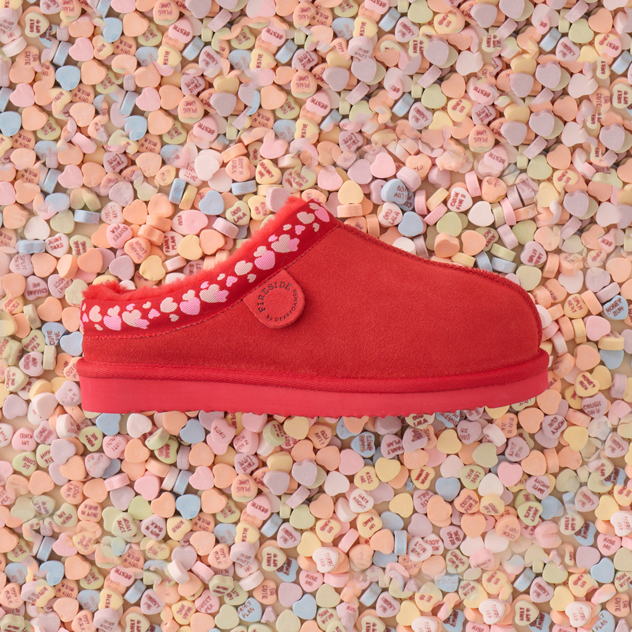 A red slipper with a decorative trim rests on a surface covered with pastel-colored heart-shaped candies.