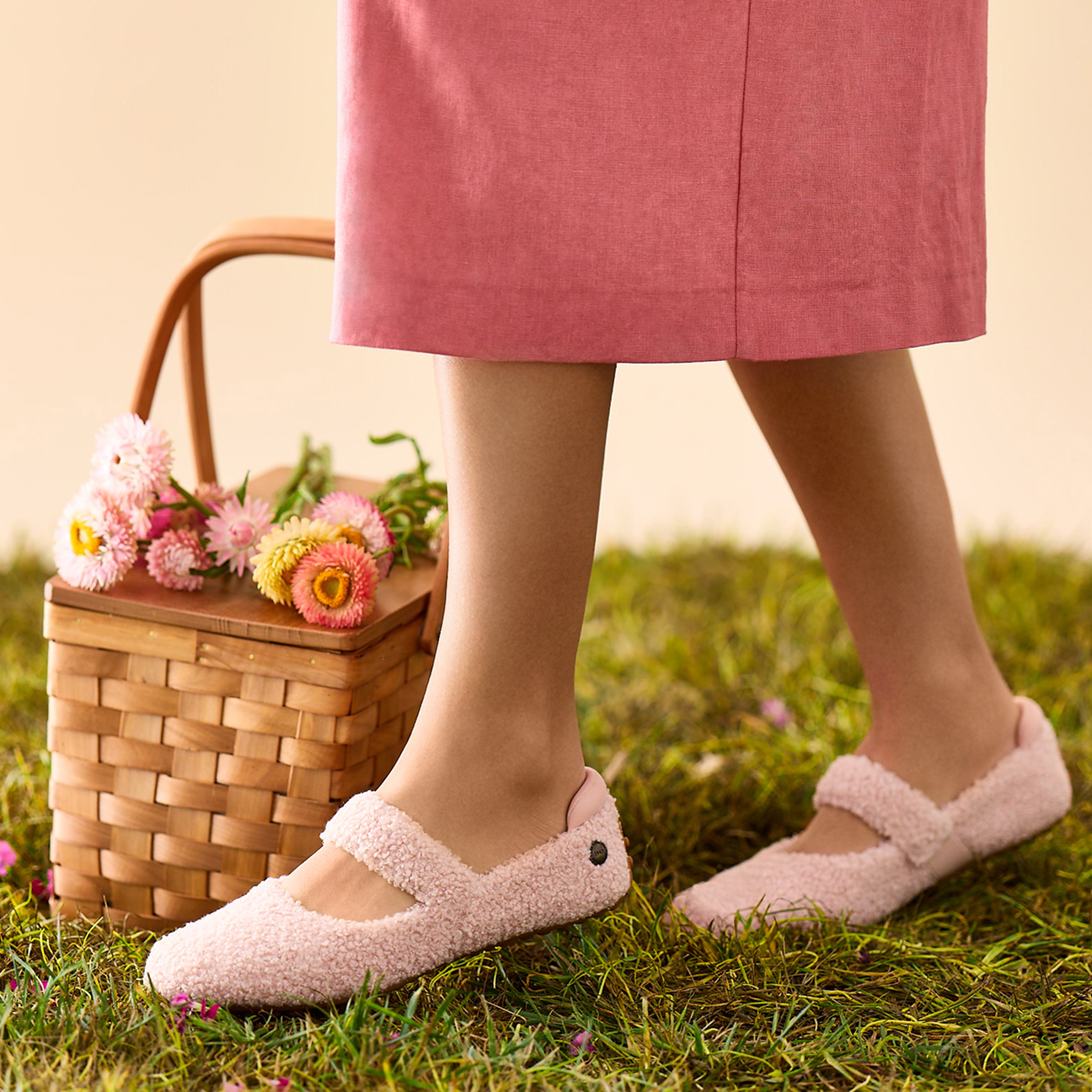 A person wearing a pink skirt and fuzzy pink shoes stands on grass next to a woven basket filled with flowers.