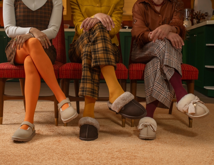 Three women seated together wearing different styles of Dearfoams Fireside styles with colorful tights and plaid pants.