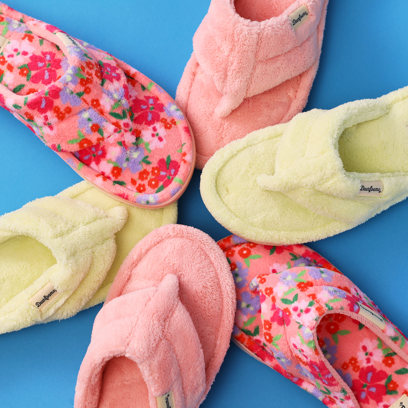 Assorted colorful plush thong slippers in floral prints, pastel pink, and soft yellow arranged on a bright blue background.