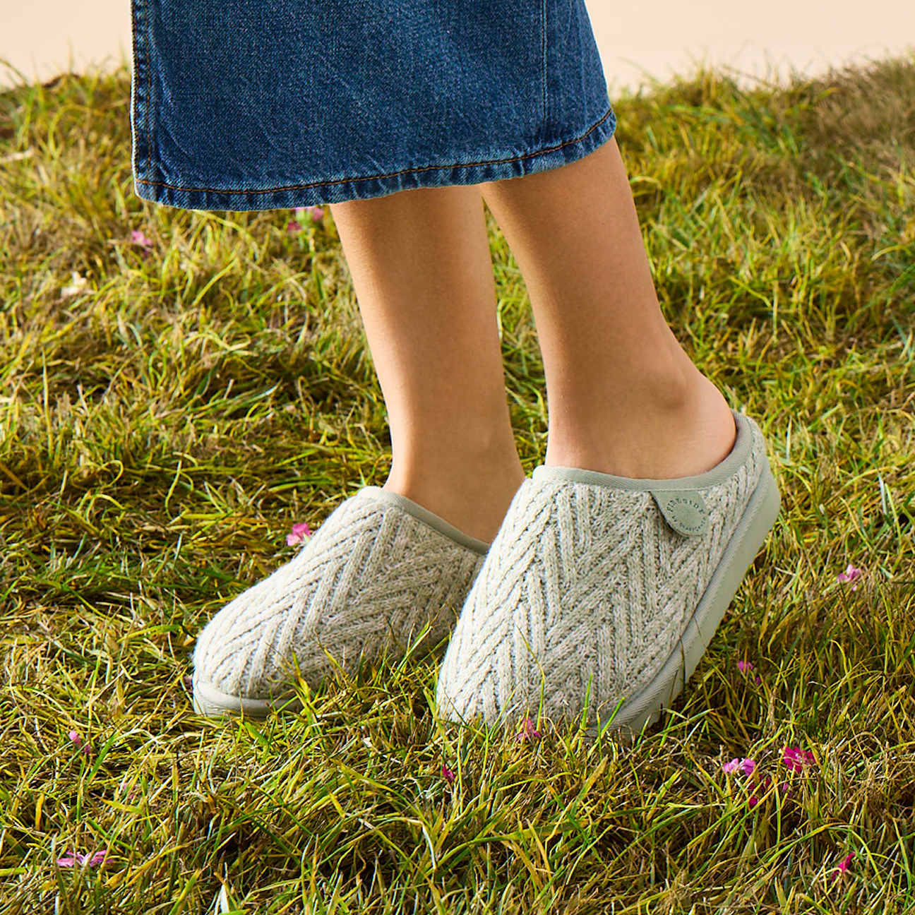 A woman wearing new light-colored, Fireside shearling slippers stands on grass in a Spring setting. Only the lower legs and slippers are visible, with a denim skirt falling just below her knees.
