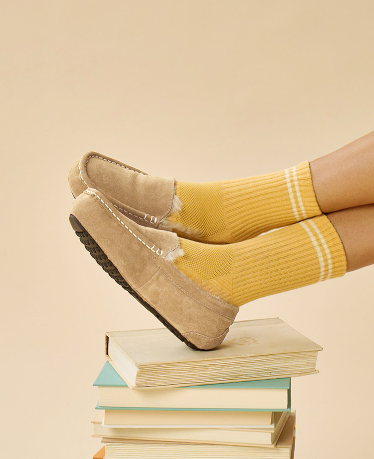 Alt text: Person wearing tan suede moccasin slippers and yellow socks while balancing on stacked books beside the Fireside by Dearfoams logo.